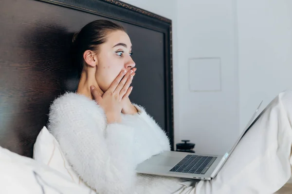 Молода здивована дівчина сидить у ліжку під білою ковдрою, дивиться жахливий фільм на своєму ноутбуці, покриває рот руками — стокове фото