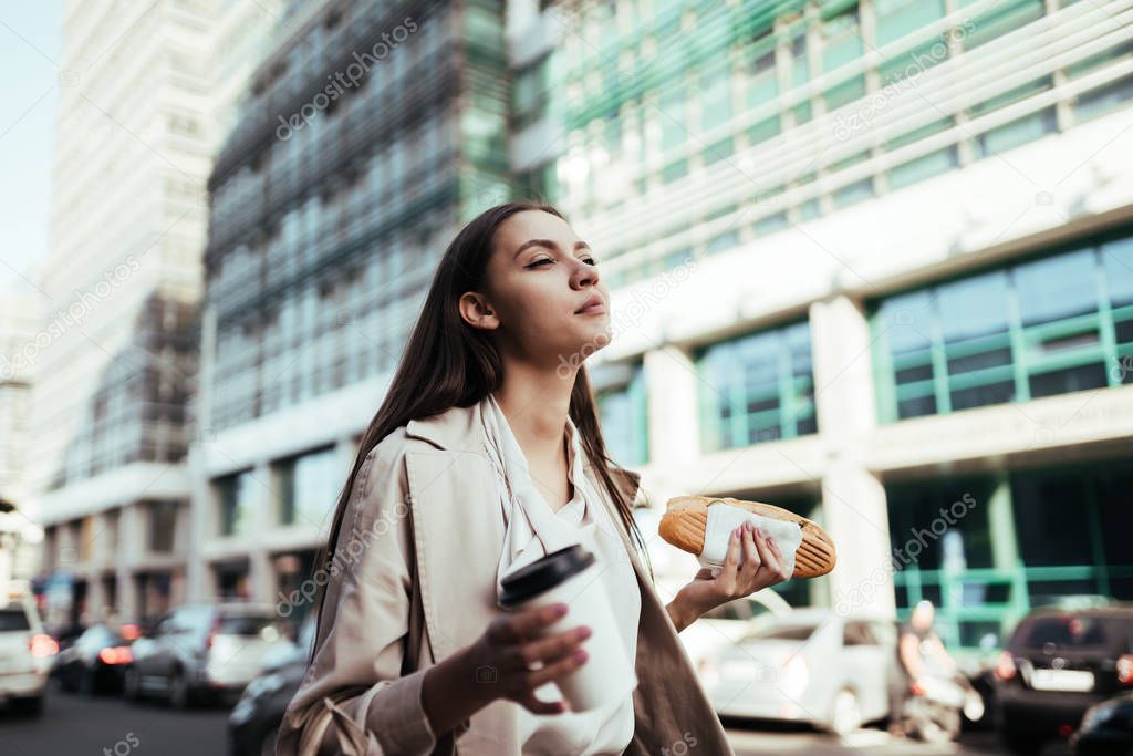 Joyful girl walks down the street and snacks on a sandwich against the background of office buildings