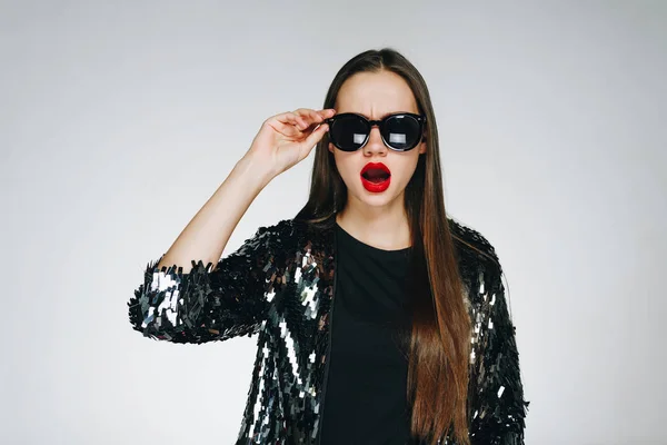 Chica de moda en una chaqueta con lentejuelas levanta gafas y se ve sorprendida . — Foto de Stock