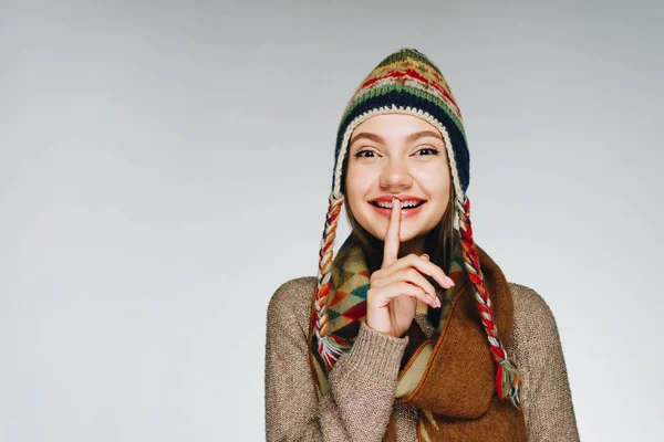 Junge fröhliche Frau mit skandinavisch warmer Mütze und Schal auf weißem Hintergrund legt den Finger an ihre Lippen und zeigt leise — Stockfoto