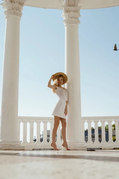 Una hermosa joven en ropa blanca y un sombrero de paja se encuentra entre columnas blancas sobre el fondo del mar — Foto de Stock