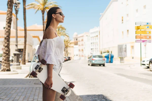 Una joven sosteniendo un monopatín en las manos de la vieja ciudad europea. Vacaciones de verano y sol. Estudiante de intercambio —  Fotos de Stock
