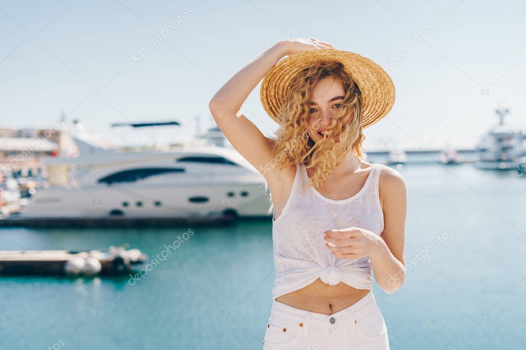 Beautiful rich girl in white clothes in a straw hat stands on the background of yachts