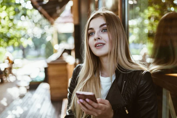 A beautiful girl in a leather jacket looks up thoughtfully, holds a phone in her hand — Stock Photo, Image
