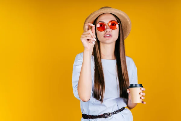 Una ragazza con un cappello di paglia e bicchieri di colore rotondo beve caffè e dice qualcosa su uno sfondo giallo . — Foto Stock