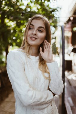 Beyaz kazaklı, uzun sarı saçlı çekici bir genç kız kendini toparlamış ve gülümseyerek yüzünü kaçırıyor.