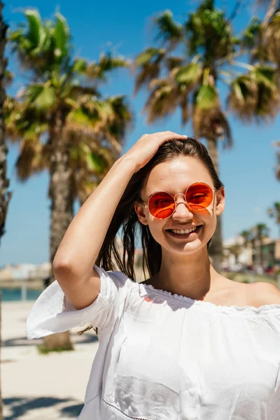 Очаровательная девушка с длинными темными волосами в белой блузке и оранжевых солнцезащитных очках улыбается и подмигивает на фоне морского побережья — стоковое фото