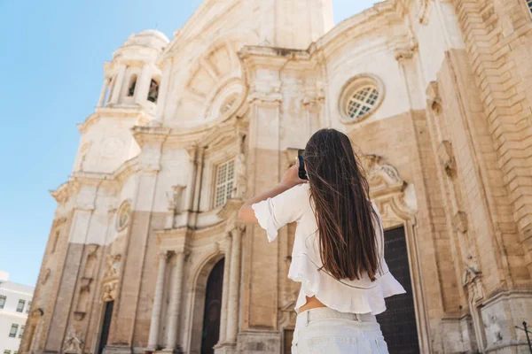 Young girl with long dark hair in a white blouse and shorts takes a photo of the cathedral on a clear day