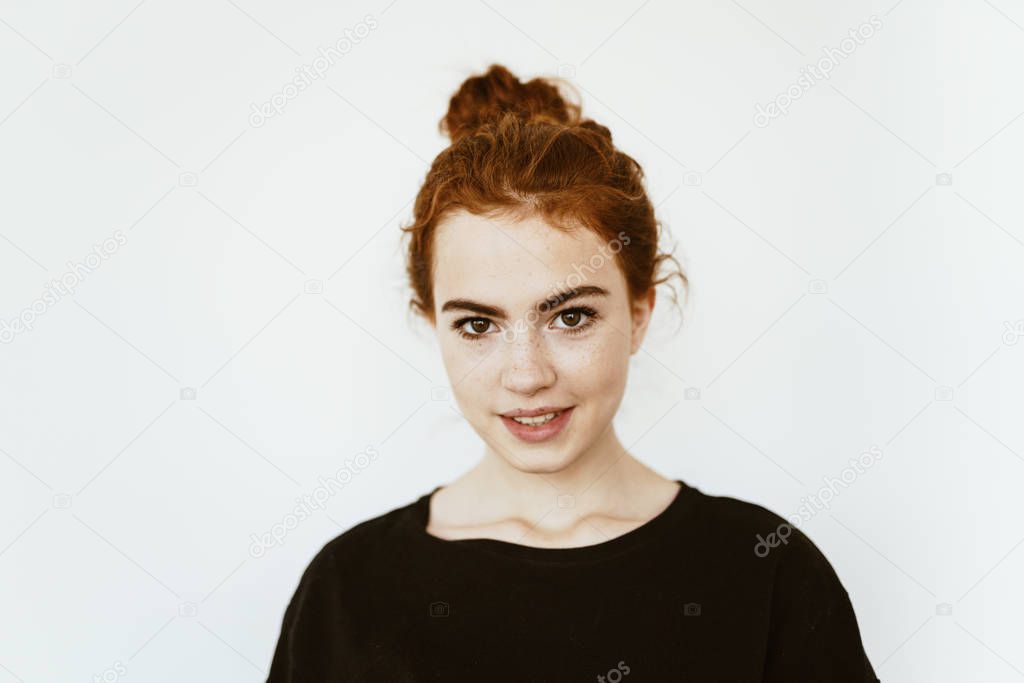 Adorable young girl with curly red hair braided in a bun on her head, with freckles on her face on a white background in a dark jacket cute smiles