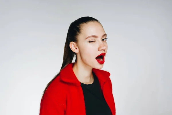Erstaunliche junge Mädchen mit dunklen langen Haaren zu einem Pferdeschwanz gebunden, mit rotem Lippenstift und in einem roten Pullover mit schwarzem Hemd auf weißem Hintergrund zwinkert mit Überraschung — Stockfoto