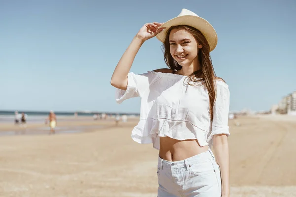 Удивительная девушка с длинными темными волосами в соломенной шляпе на голове в белой футболке и шортах улыбается на песчаном берегу в солнечный день — стоковое фото