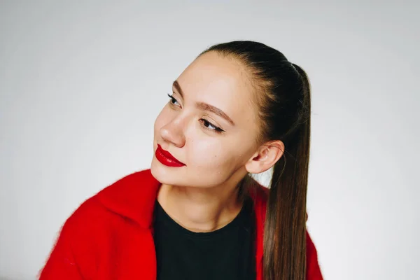 Menina incrível com cabelo longo escuro amarrado em um rabo de cavalo, com batom vermelho e em uma camisola vermelha com uma camiseta preta em um fundo branco, examina algo de lado com interesse — Fotografia de Stock