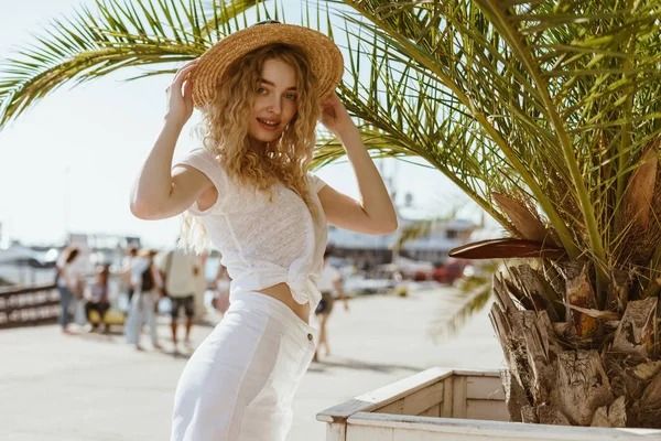 Ελκυστική νεαρή κοπέλα με μακριά σγουρά μαλλιά στο λευκό T-shirt και φούστα, κρατά ένα ψάθινο καπέλο στο κεφάλι της και στέκεται στο φόντο της με θέα στη θάλασσα — Φωτογραφία Αρχείου