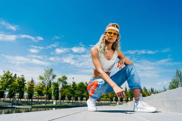Sportief gebruind meisje met lang blond krullend haar in de witte kant top en blauwe joggingbroek met een gele kap zit op de achtergrond van het gazon en kijkt naar de zijkant. — Stockfoto