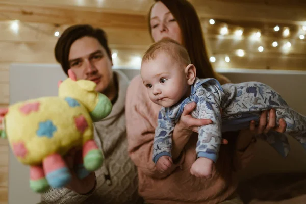 Joven familia de mamá y papá con su hijo están sentados en el sofá, donde mamá tiene un hijo pequeño y papá le muestra un juguete suave — Foto de Stock