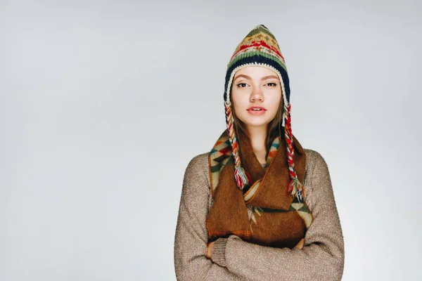 Attraktives junges Mädchen mit dunklen langen Haaren mit Hut im skandinavischen Stil und braunem Schal, bekleidet mit einem braunen Pullover, umarmt sich auf weißem Hintergrund und sieht nachdenklich aus — Stockfoto