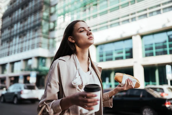 Giovane ragazza d'affari in cappotto beige mangia in movimento, tenendo un rotolo nella mano destra e un bicchiere di carta nella mano sinistra, correndo a una riunione — Foto Stock