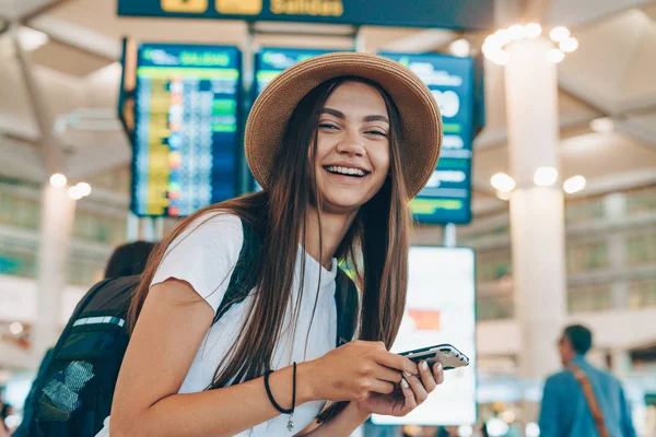 Tanned girl with long dark hair in a straw hat on her head and the white T-shirt with  phone in her hands and backpack on her back is looking forward to her flight at the airport to the plane — Stock Photo, Image