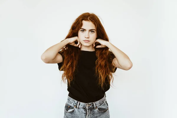 Nettes junges Mädchen mit langen lockigen roten Haaren mit Sommersprossen im Gesicht im schwarzen T-Shirt und Jeans auf weißem Hintergrund glättet ihr Haar — Stockfoto