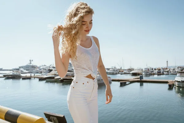 Lockigt blond står på parapet vid havet, stänga ögonen med glädje mot bakgrund av fartyg — Stockfoto