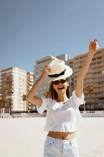 Alegre chica en la playa exclama en sorpresa, con energía levantando su sombrero y haciendo un gesto de victoria con la otra mano — Foto de Stock