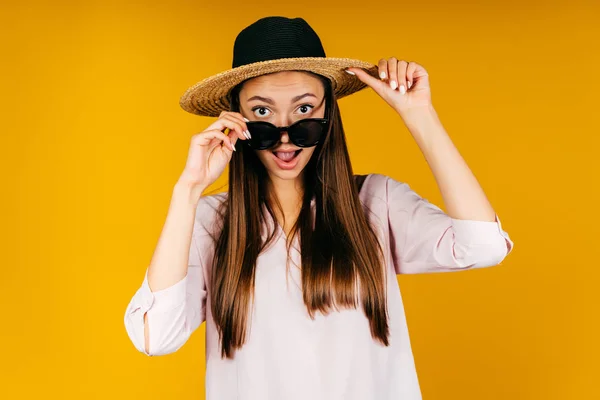 Fondo amarillo. chica abre la boca con su lengua y mira por encima de las gafas en un sombrero — Foto de Stock