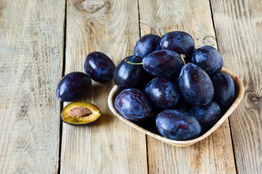 Su ile olgun mavi Erik ahşap arka plan üzerinde ahşap kase geliyor. Yarım bir Erik bir taşla. Yaz mevsim meyveleri kavramı. Rustik tarzı.