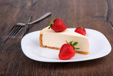 Çilekli cheesecake dilimi beyaz bir tabakta ahşap arka planda. Arka plandaki çatal bulanık. Yumuşak odak.