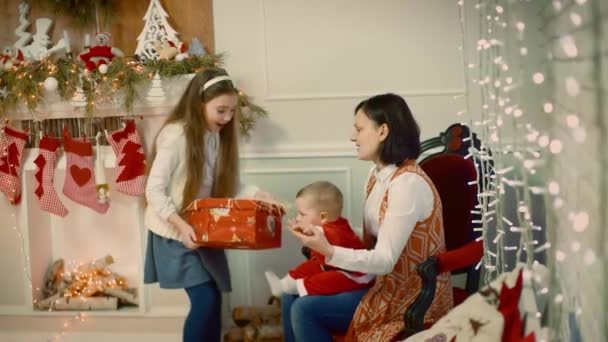 Increíble mirada de una niña le da a un niño un regalo a un niño que se sienta en su regazo de mamás cerca de un árbol de Navidad decorado y regalo . — Vídeos de Stock