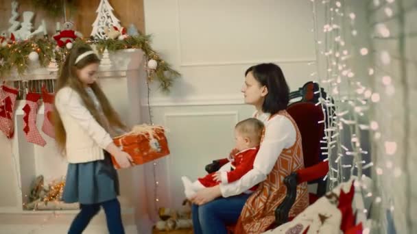 Increíble mirada de una niña le da a un niño un regalo a un niño que se sienta en el regazo de su madre y sonríe cerca de un árbol de Navidad decorado y regalo . — Vídeos de Stock