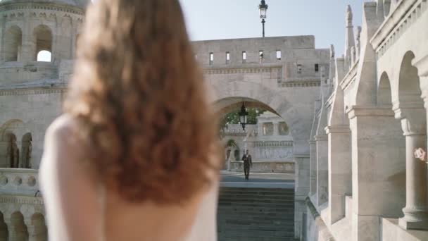 Μια πολύ όμορφη νεαρή γυναίκα ή νύφη σε ένα λευκό φόρεμα, μείνετε στο μπαλκόνι του παλιού κάστρου, και εξετάζει το πώς ο γαμπρός πηγαίνει σε αυτήν στην πόλη Βουδαπέστη — Αρχείο Βίντεο