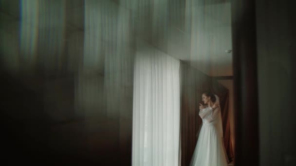 Молодая красивая невеста останавливается у окна и ждет жениха и смотрит в окно — стоковое видео