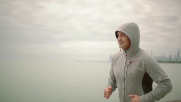 Een jonge sportieve man loopt tegen de achtergrond van de stad en het meer, langzame motie — Stockvideo