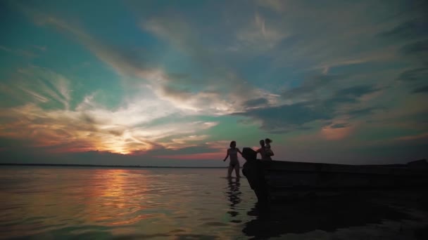 A mulher e o homem dão as mãos e correm alegremente ao longo do lago ao pôr-do-sol. As silhuetas de umas crianças pequenas e os pais juntam-se a seguir, correm e brincam. Movimento lento . — Vídeo de Stock