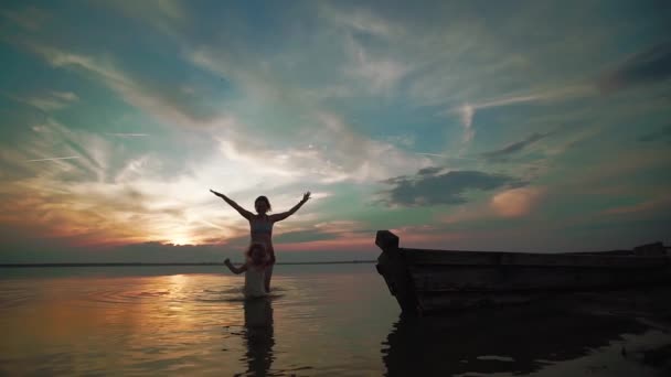 Zeitlupe. Eine Frau und ein kleines Kind laufen bei Sonnenuntergang fröhlich auf dem Wasser eines Sees. Silhouetten von kleinen Kindern und Eltern, die nebeneinander laufen und spielen. — Stockvideo