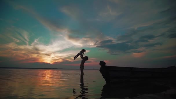 Slow Motion. Een vrouw heft haar baby op uit het meer en draait met haar vrolijk rond met haar op een meer bij zonsondergang. Silhouetten van een kleine kinderen en ouders tijd samen volgende, rennen en spelen. — Stockvideo