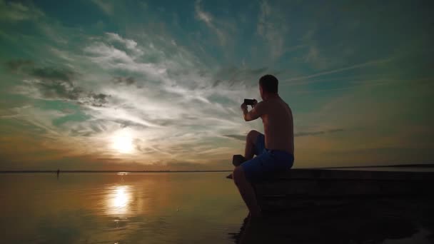 Slow Motion. Een man zit op een boot en neemt een foto op de telefoon van een ongelooflijke zonsondergang een meer bij zonsondergang — Stockvideo