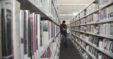 Üniversite öğrencisi kütüphanede yerde duruyor, kitap okuyor. Dikey şekil, yan görünüm, tam uzunluk,