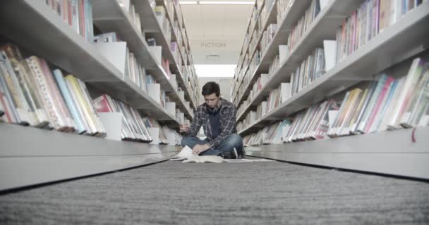 Studentka siedzi na podłodze w bibliotece, czytając książkę. Zamienia strony księgi — Wideo stockowe