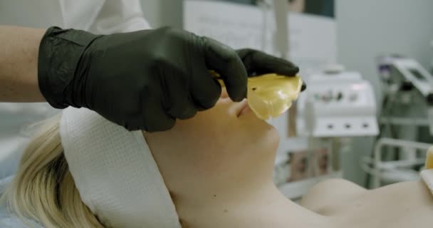 Kosmetikerin trägt goldene Gesichtsmaske auf das Gesicht der Frau auf — Stockvideo