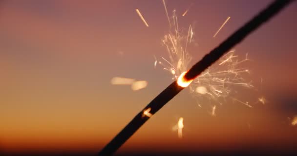 Uma varinha ardente brilhante em câmera lenta de fogo bengali contra o fundo do céu incrível bokeh ao pôr do sol do dia. Luzes laranja caem de todos os lados. Padrão de Natal e Feliz Ano Novo. Fechar — Vídeo de Stock