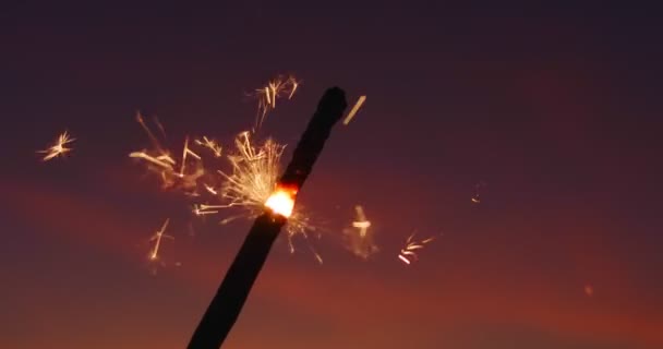 Una varita ardiente brillante de fuego bengalí en cámara lenta contra el fondo del asombroso cielo bokeh al atardecer del día. Las luces naranjas caen por todos lados. Patrón de Navidad y Feliz Año Nuevo. De cerca. — Vídeos de Stock