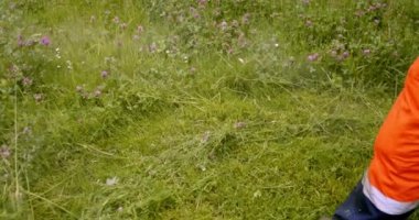 Turuncu tulumlu işçi çimleri biçer çim biçme makinesiyle, yavaş çekimde arkadan bakar.