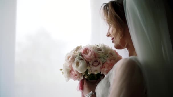 La mañana de la novia de pie junto a la ventana en un peignoir blanco sostiene en sus manos un ramo de rosas de boda. .. De cerca. Mover cámara — Vídeos de Stock