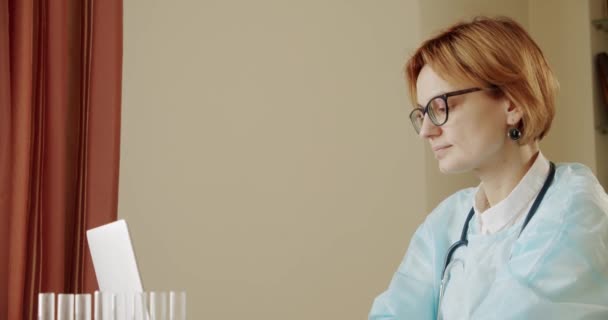 Doctora sentada en el escritorio trabajando en la computadora portátil. En cámara lenta. Tiro medio. — Vídeos de Stock