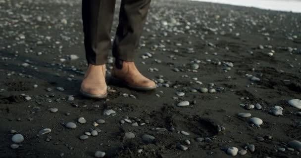 Μια κοντινή θέα των ποδιών ενός άνδρα σε καφέ μπότες με τα πόδια σε μαύρη άμμο και πέτρες σε μια παραλία στην Ισλανδία. — Αρχείο Βίντεο
