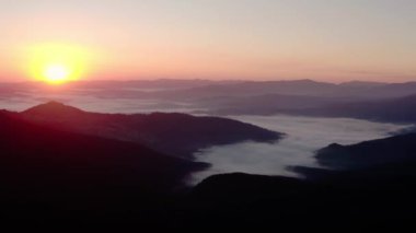 Hava aracı görüntüsü. Dağlarda gün doğumu veya günbatımı, güneşin altında ağaçların üzerinden uçmak.