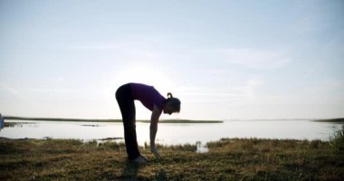 Arka planda gökyüzü ve gölde jimnastik yapan genç bir kadının silueti. V2