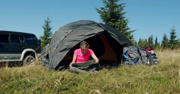 Młody człowiek zwiedzający mapę siedzącą w pobliżu namiotu i samochodu w górach w słoneczny dzień. — Wideo stockowe