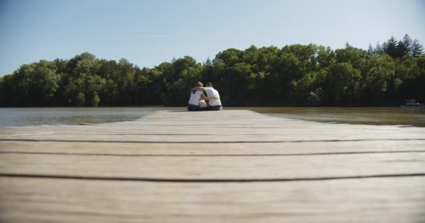 Familj mamma baby och make sitter på en träpir vid dammen. — Stockvideo